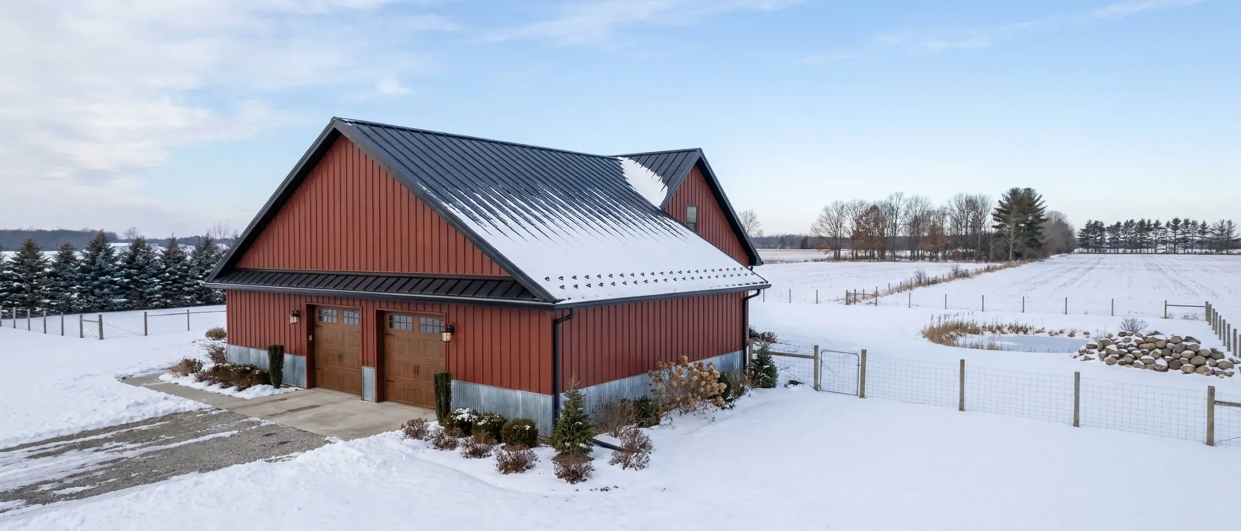 A completed barndominium in winter viewed from a low angle, showing its standing seam roof with snow guards, wall panels, gutters and wall lights.